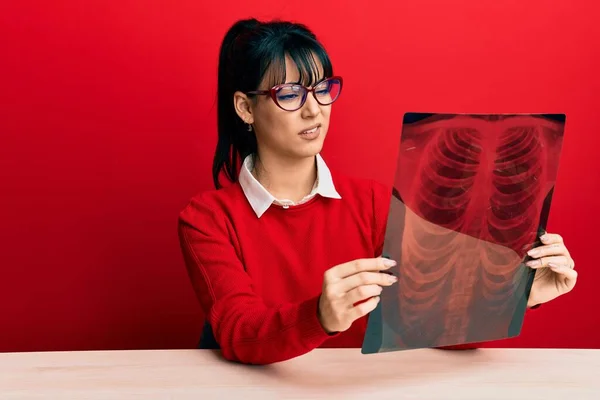 Young brunette woman with bangs holding chest radiography clueless and confused expression. doubt concept.