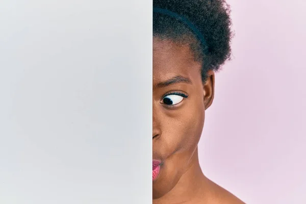 Young african american girl holding blank empty banner over half face making fish face with mouth and squinting eyes, crazy and comical.