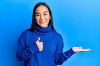 Genç Asyalı kadın, günlük kışlık süveter giyiyor avuç içi gösteriyor ve baş parmaklarıyla iyi bir jest yapıyor, mutlu ve neşeli gülümsüyor. 