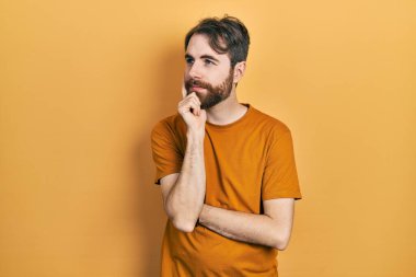 Sakallı, sıradan sarı tişörtlü, eli çenesinde soru soran beyaz bir adam. Düşünceli bir ifade. Gülümseyen ve düşünceli bir yüz. şüphe kavramı. 