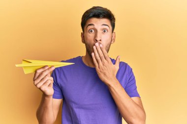 Elinde kağıt uçak tutan, ağzı eliyle kapatan genç ve yakışıklı bir adam. Şok olmuş ve hata yapmaktan korkuyor. şaşırmış ifade 