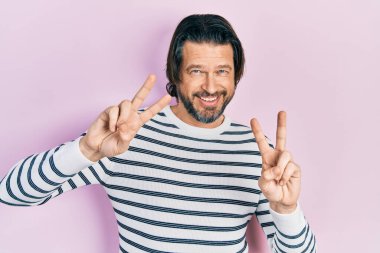 Orta yaşlı, günlük kıyafetler giyen, gülümseyen, parmaklarını zafer işareti yaparken gösteren beyaz bir adam. İki numara.. 