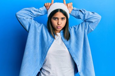 Şirin yün şapkalı genç Latin kadın komik jestler yapıyor ve parmağıyla boynuz yapıyor. 