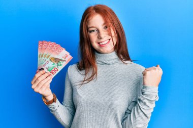 Genç kızıl saçlı kız elinde 100 yeni Zelanda doları banknotuyla gururla bağırıyor, zaferi ve başarıyı kutluyor. 