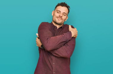 Gündelik kıyafetler giyen genç İspanyol adam kendine sarılıyor mutlu ve pozitif, kendinden emin gülümsüyor. Sevgi ve özgüven. 