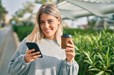 Genç sarışın kız akıllı telefon kullanıyor ve şehirde kahve içiyor.