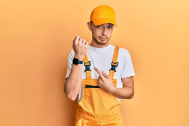 Tamirci üniforması giyen İspanyol genç adam acele ederek saati, sabırsızlığı ve rahat bir ifadeyle kameraya bakıyor. 