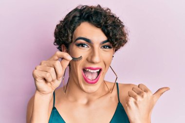 Sahte kirpiklerle makyaj yapan genç bir adam baş parmağını kaldırıyor ve ağzı açık gülümsüyor. 