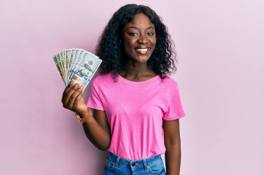 Dolarları tutan güzel Afrikalı genç bir kadın. Pozitif ve mutlu görünüyor. Duruyor ve gülümsüyor. 