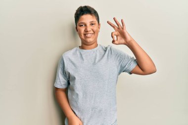 Gündelik gri tişört giyen genç İspanyol çocuk gülümsüyor ve el ve parmaklarıyla tamam işareti yapıyor. başarılı ifade. 