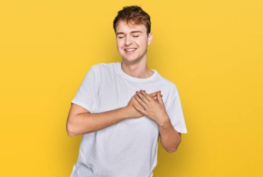 Gündelik beyaz tişört giyen, elleri göğsünde, gözleri kapalı gülümseyen ve yüzünde minnet ifadesi olan genç beyaz bir adam. sağlık kavramı. 