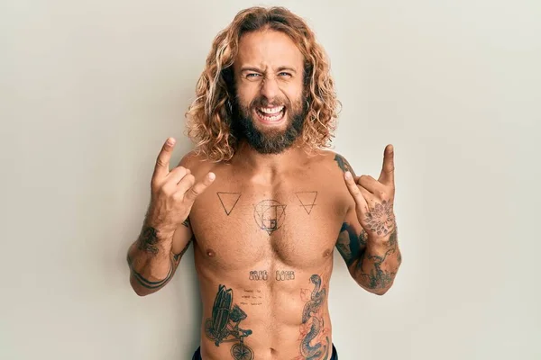 Handsome man with beard and long hair standing shirtless showing tattoos shouting with crazy expression doing rock symbol with hands up. music star. heavy concept.
