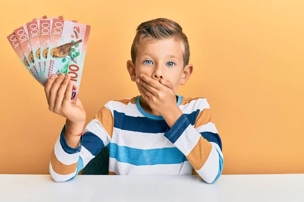 手で口をカバーテーブルの上に座っている100の新しいニュージーランドドル紙幣を保持している愛らしい白人の子供はショックを受け 間違いを恐れています 驚きの表情 — ストック写真