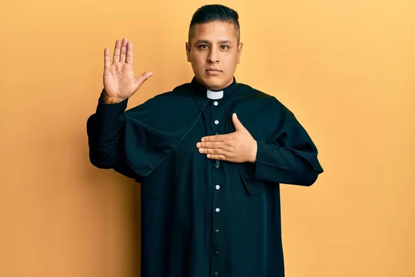 胸と手のひらに手で身に着けている黄色の背景を身に着けている若いラテンの司祭の男 忠誠の誓いを作る — ストック写真