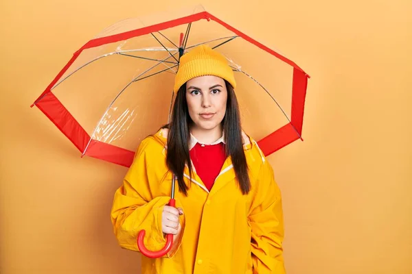 Junge Hispanische Frau Gelbem Regenmantel Mit Regenschirmdenken Und Nüchternem Gesichtsausdruck — Stockfoto
