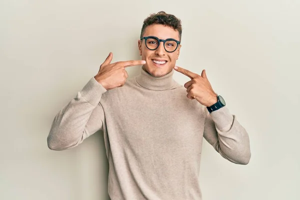 カジュアルなタートルネックセーターを身に着けているヒスパニック系の若者は 陽気な笑みを浮かべて指の歯と口を指しています 歯の健康の概念 — ストック写真