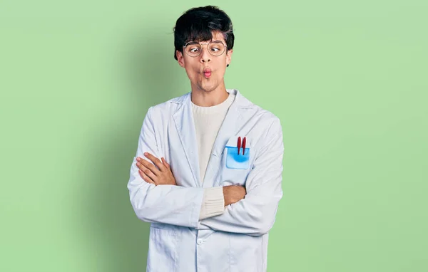 Handsome hipster young man with crossed arms wearing doctor uniform making fish face with mouth and squinting eyes, crazy and comical.