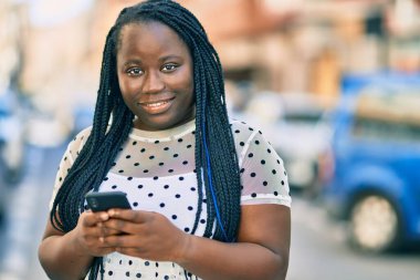 Genç Afrikalı Amerikalı kadın şehirde akıllı telefon kullanarak mutlu bir şekilde gülümsüyor..