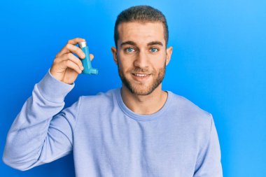 Elinde astım spreyi tutan genç beyaz bir adam. Pozitif ve mutlu görünüyor. Kendine güvenen bir gülümsemeyle dişlerini gösteriyor. 