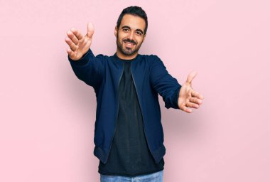 Gündelik giysiler giymiş genç İspanyol adam kameraya bakıp sarılmak için kollarını açmış gülümsüyor. Mutluluğu kucaklayan neşeli bir ifade.. 