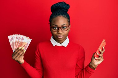 Genç Afrikalı Amerikalı kadın elinde Norveç Kronu banknotları tutan akıllı telefon kullanıyor. Yüzünde ciddi bir ifadeyle rahatlamış. Basit ve doğal bir şekilde kameraya bakıyorsun.. 