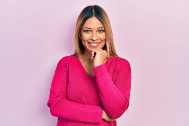 Güzel İspanyol kadın, sıradan pembe bir süveter giyip kameraya kollarını kavuşturup, çenesine dokunarak gülümsüyor. Olumlu düşünmek. 