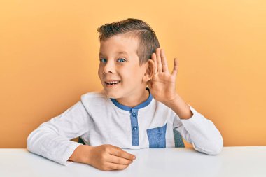 Kafkasyalı sevimli bir çocuk, günlük giysiler içinde masada oturmuş, sırıtarak dedikodu ve dedikoduları dinliyor. Sağırlık kavramı. 