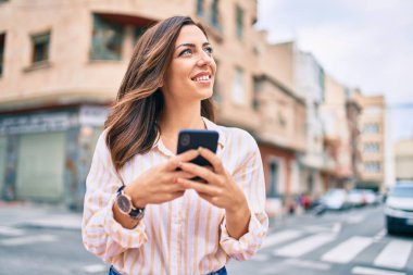 Genç İspanyol kadın şehirde akıllı telefon kullanarak mutlu bir şekilde gülümsüyor..
