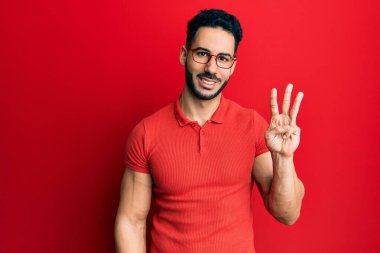 Gündelik kıyafetler giyen ve gözlük takan İspanyol genç adam üç numarayı gösterip işaret ederken kendinden emin ve mutlu bir şekilde gülümsüyor.. 