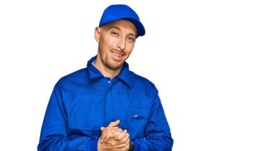 Kel, sakallı, işçi tulumu giyen, elleri birbirine dolanmış ve gülümseyen, gevşemiş ve neşeli elleri çapraz parmaklı adam. Başarı ve iyimserlik 