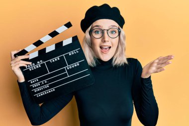 Genç sarışın kız elinde video film panosu ile başarıyı mutlu bir gülümsemeyle kutluyor ve kazanan el kaldırarak ifade ediyor. 