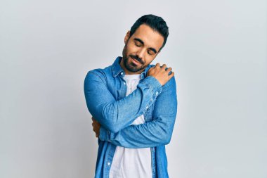 Gündelik kıyafetler giyen genç İspanyol adam kendine sarılıyor mutlu ve pozitif, kendinden emin gülümsüyor. Sevgi ve özgüven. 