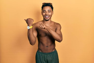 Mayo ve yüzme gözlüğü takan genç Afro-Amerikalı adam, arka tarafı işaret ediyor. El ve baş parmakları havada, kendine güvenen. 