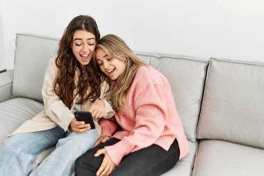 Genç çift evde koltukta oturmuş akıllı telefon kullanarak mutlu bir şekilde gülümsüyor..