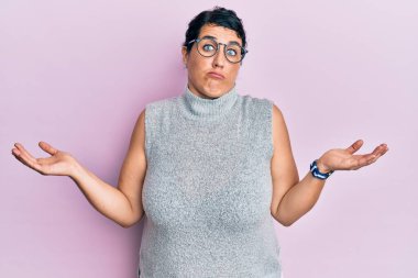 Artı, sıradan kıyafetler giyen, gözlüklü, kafası karışmış, elleri ve kolları havada esmer bir kadın. şüphe kavramı. 