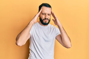 Sakallı, sıradan beyaz tişörtlü, baş ağrısı çeken, çaresiz ve stresli bir genç adam. Ağrı ve migren yüzünden. Eller başının üstünde.. 