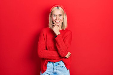 Güzel sarışın kadın, sıradan kırmızı bir kazak giyiyor kameraya bakıyor kollarını kavuşturup gülümsüyor ve elini çenesine kaldırıyor. Olumlu düşünmek. 