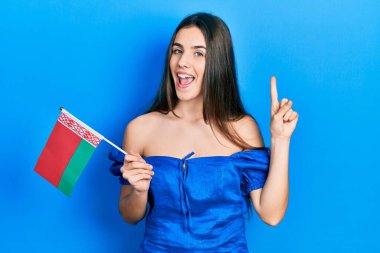 Genç esmer genç elinde Belarus bayrağı tutarken bir fikir ya da soruyla gülümsüyor ve mutlu bir yüzle işaret ediyor. 