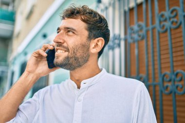 Güneşli bir günde sakallı, günlük beyaz gömlek giyen yakışıklı adam dışarıda mutlu bir şekilde gülümserken akıllı telefon kullanıyor.