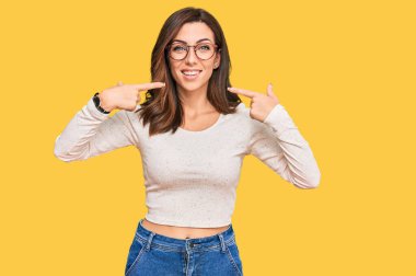 Genç esmer kadın gündelik kıyafetler ve gözlük takıyor neşeli bir şekilde gülümsüyor ve parmaklarıyla ve ağzıyla işaret ediyor. diş sağlığı kavramı. 