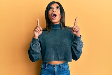 Gündelik giysiler giyen genç Latin transseksüel kadın şaşkın ve şaşırmış bir şekilde yukarı bakıyor ve parmaklarıyla işaret ediyor ve kollarını kaldırıyor.. 