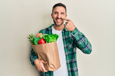 Sakallı yakışıklı bir adam, elinde torbayla elinde alışveriş torbalarıyla, parmakları burnuna ve yüzüne dönük, neşeli bir şekilde gülümsüyor. güzellik konsepti 