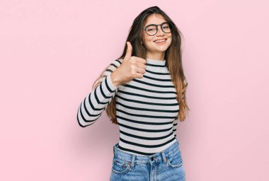 Gündelik giysiler ve gözlük takan genç ve güzel bir kız mutlu bir el hareketi yapıyor. Kameraya bakıp başarıyı gösteren ifade. 