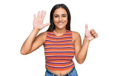 Genç esmer kadın gündelik kıyafetler giyip altı numaralı parmaklarıyla işaret ederken kendinden emin ve mutlu bir şekilde gülümsüyor.. 