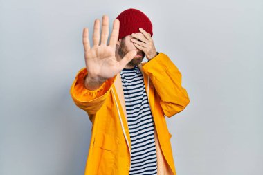 Sakallı, sarı yağmurluk giyen, elleriyle gözlerini kapatan, üzgün ve korku ifadeli beyaz bir adam. utangaç ve olumsuz kavram. 