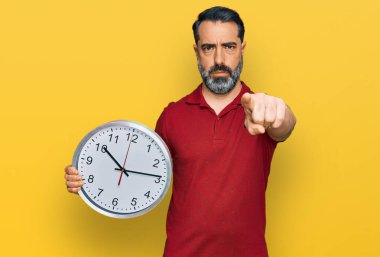 Orta yaşlı, sakallı, büyük saati tutan, kamerayı işaret eden bir adam. Kendine güvenen, ciddi görünen bir jest. 