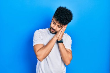 Sakallı, beyaz tişörtlü, uykulu genç bir Arap adam. Yorgun, hayal kuruyor, el ele poz veriyor ve gözleri kapalı gülümsüyor.. 