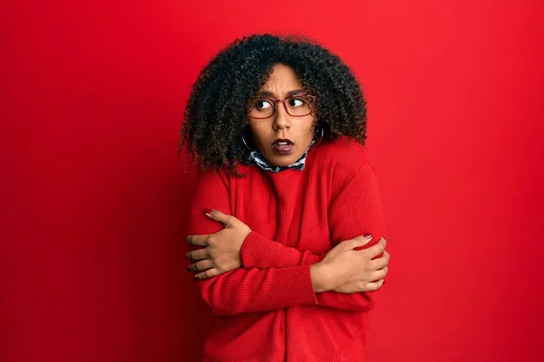 Beautiful african american woman with afro hair wearing sweater and glasses shaking and freezing for winter cold with sad and shock expression on face