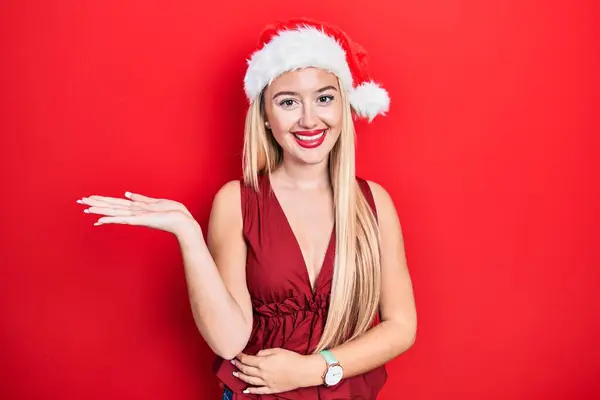 Молодая Блондинка Рождественской Шляпе Улыбается Веселым Подарком Указывает Ладонью Камеру — стоковое фото