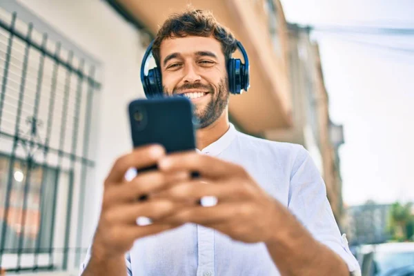 晴れた日に白いシャツを着て髭を生やしたハンサムな男は ヘッドフォンを着て音楽を聞いてスマートフォンを使って屋外で幸せな笑顔 — ストック写真